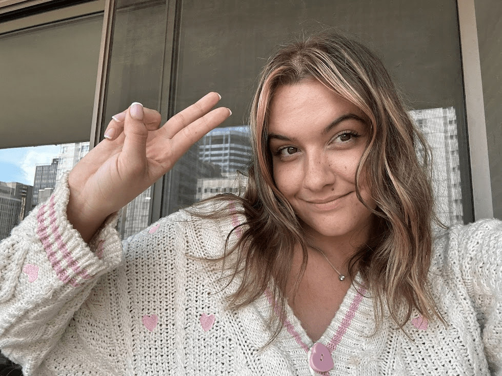 Sydney wearing a white sweater with pink hearts, and she is smiling and making a peace sign with her hand. The background features city buildings and a balcony.